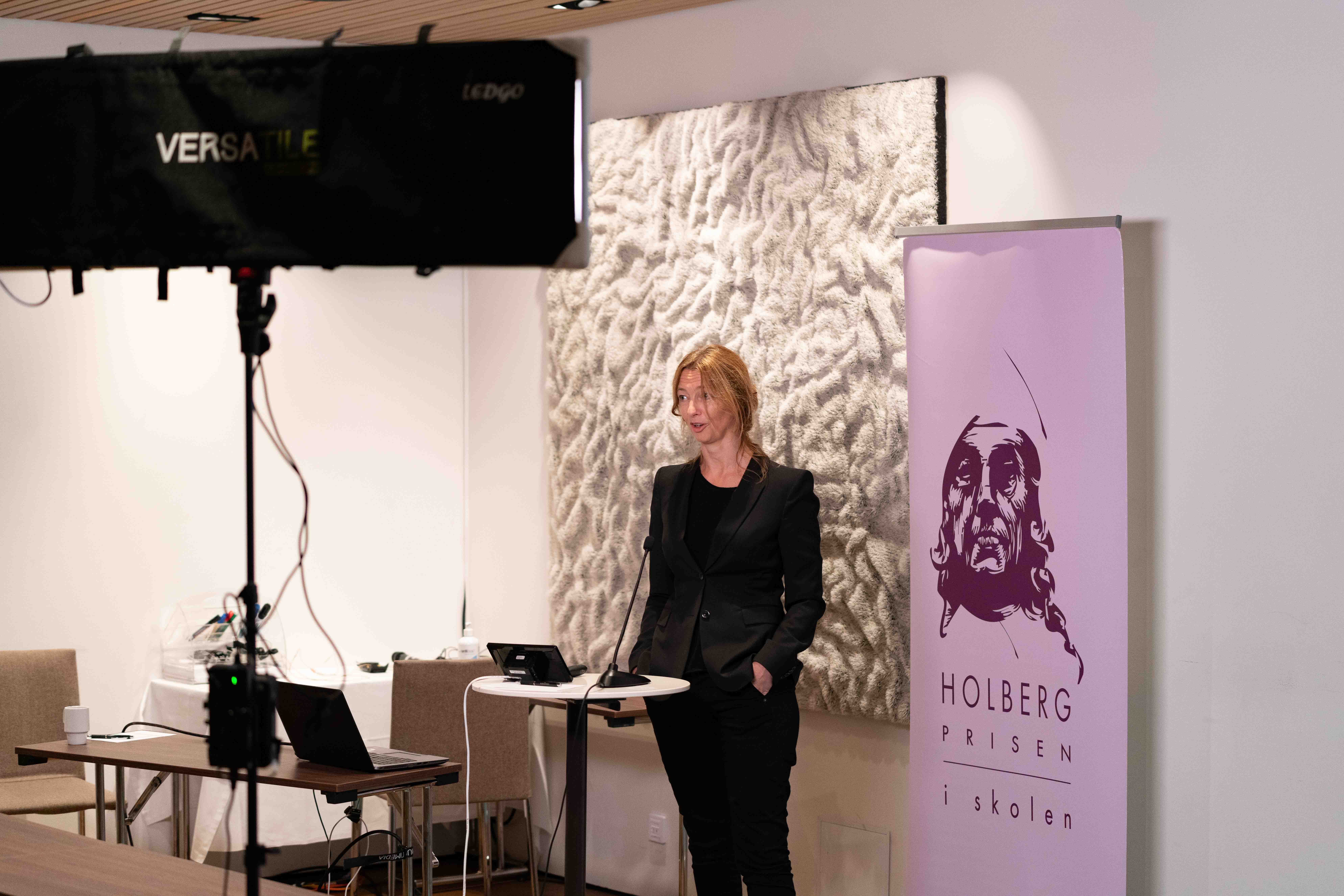Bilde av fagkonsulent Marie von der Lippe stående i oppsettet for digital avvikling: Foran en roll up med Holbergprisen i skolens logo, og et bord med mikrofon, og bak en lyskaster. Foto: Thor Brødreskift